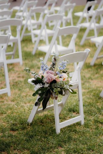 A vertical shot of a white chair with a floral bouquet and other white chairs at an outdoor wedding