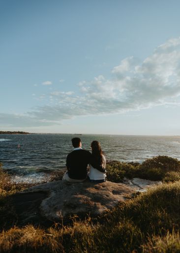 A back view of a young couple sitting on a rock and admiring the view of the sea