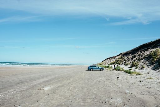 A car parked at the beach surrounded by the sea under the sunlight and a blue cloudy sky