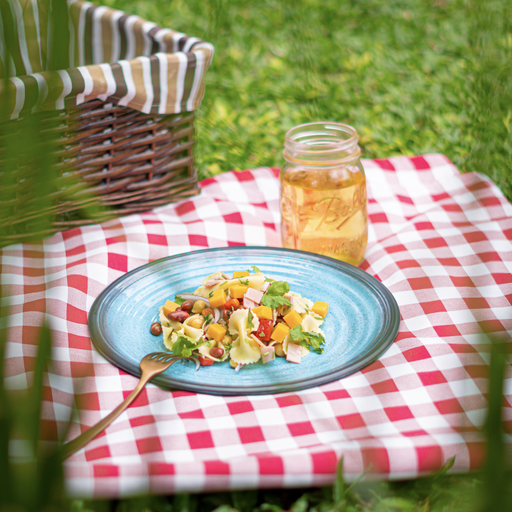 A closeup of the salad with macaroni, cheese, and other colorful ingredients on a plate next to a jar of juice and a straw basket on a picnic blanket