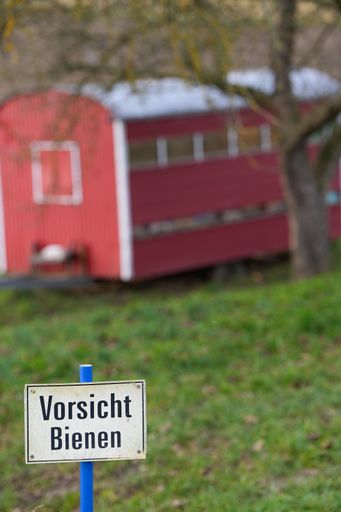 A vertical shot of a red beehouse with a sign in front saying "be aware of bees" in German