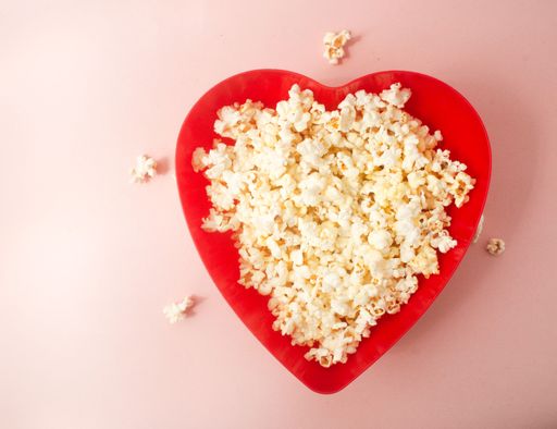 A red heart-shaped box filled with popcorn on a pink background