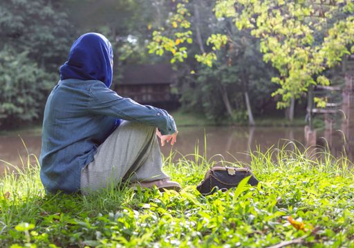A person with a jacket and a hood sitting on green grass against a lake on a sunny day