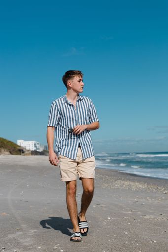A young handsome Caucasian boy in a short sleeve shirt with stripes and shorts walking on the beach