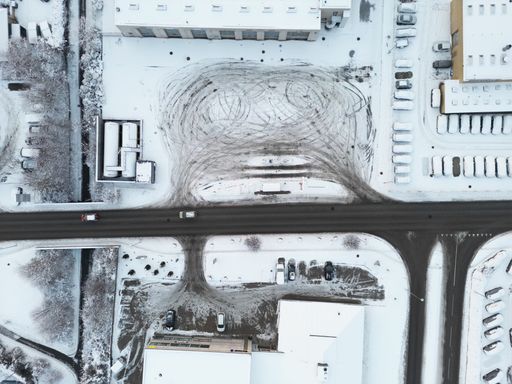 An aerial top view of a road crossing through a snow-covered  area with buildings, parked cars and wheel trails on snow