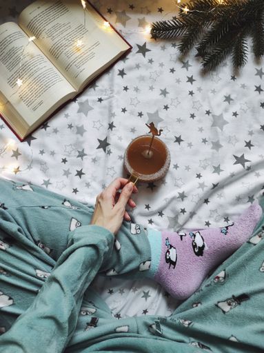 A vertical top view of a woman hand with a cup of tea on a soft bed with a book and Christmas lights.