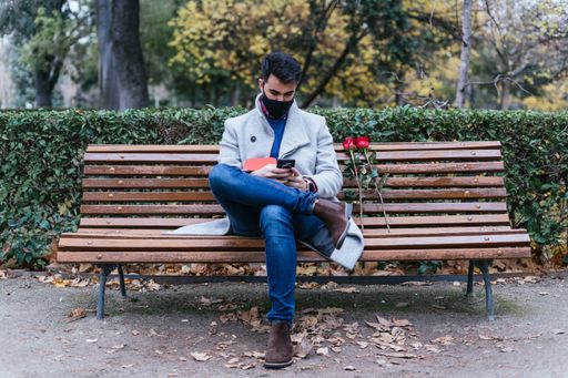A closeup of a male holding a mobile phone with red roses beside him on a bench outdoors-concept Valentine's day