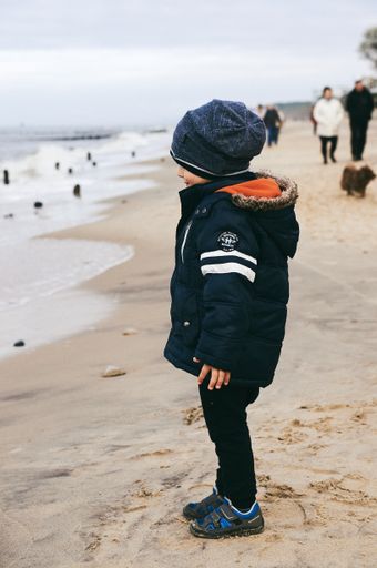 Young boy standing on sand close by the water at a beach on a cold day.
