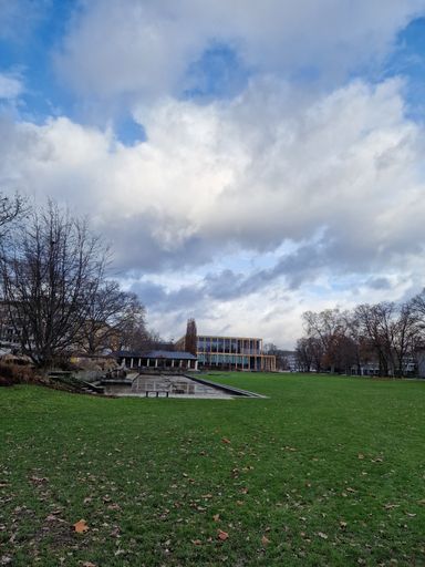 A vertical shot of buildings in the distance and a green park under the blue sky and clouds