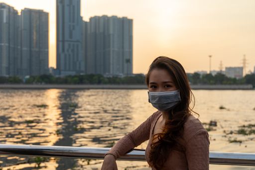 An attractive Asian female in a facial mask posing in front of a lake and skyscrapers during sunset - new normal concept