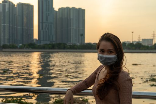 An attractive Asian female in a facial mask posing in front of a lake and skyscrapers during sunset - new normal concept