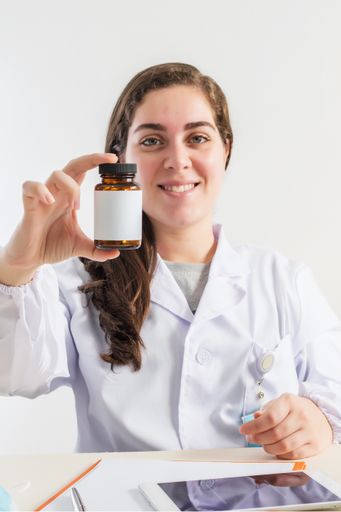 A vertical shot of a smiling female doctor holding a bottle while sitting in her consulting room