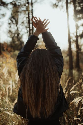 A vertical shot of a young woman standing in a lush, green forest, her arms raised up towards the sky in an expression of joy and freedom