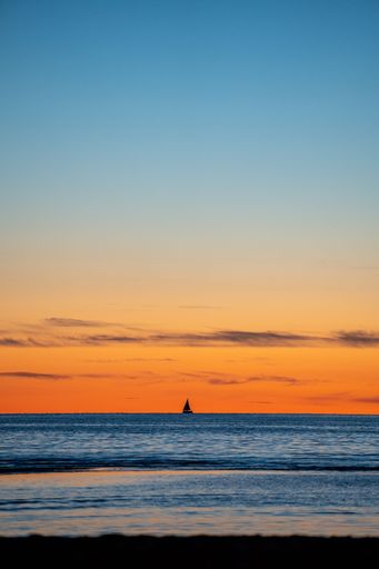 A vertical shot of a single sailing boat in the distance against the orange sky at sunset