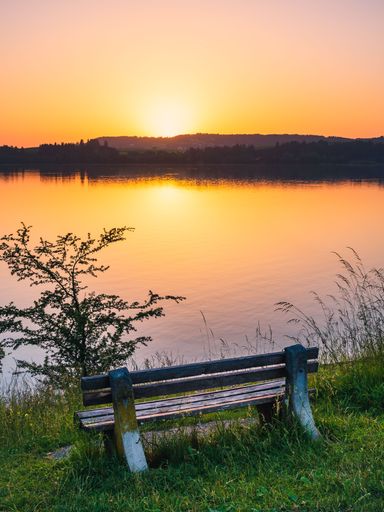 A vertical shot of a wooden bench on the grass at a lakeside during sunset with orange sky