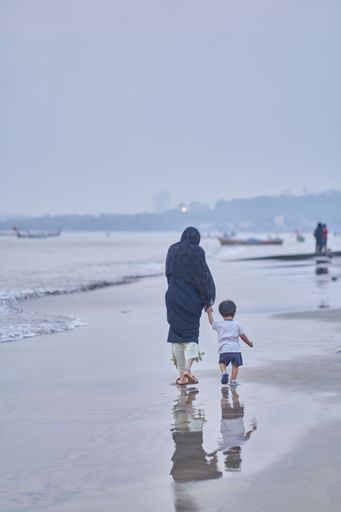 A vertical shot of a mother holding her child's hand as they walk on the shore of a beach