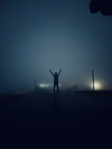 A vertical shot of a silhouette of a joyful person showing peace signs on a foggy evening