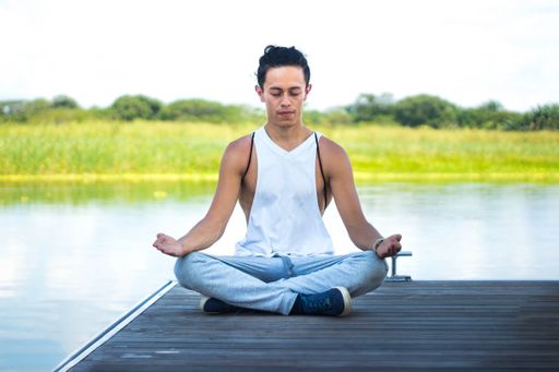 A handsome young man in a ponytail and a white t-shirt meditating in front of the water and the field