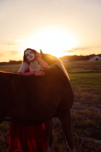 A beautiful woman in a red dress posing in a meadow with a horse at sunset.
