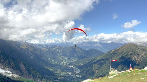 An aerial view of a paraglider flying above lush green mountain range and valleys under blue cloudy sky