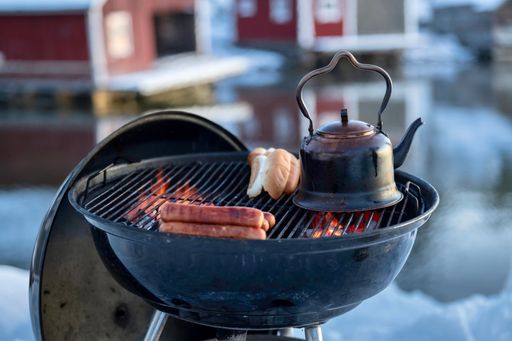 Grilling grid with sausages, bread, and teapot situated on the edge of a frozen lake in wintertime
