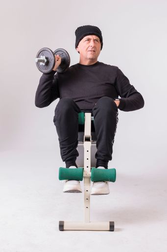 A Caucasian man sitting on the sit up excerside machine and doing strength training exercise with a barbell against a white background
