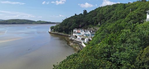A drone shot of a building on the seaside with a cliff covered with greenery under blue cloudy sky