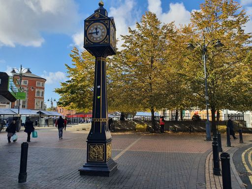 A clock on the street with walking people nearby.