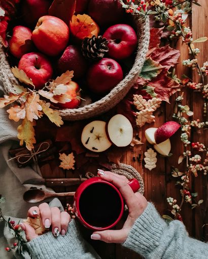 A vertical top view of a woman holding a cup of coffee with a basket full of apples on a wooden table