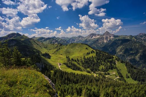 A bird's eye view of a mountain range covered with green forests under a cloudy blue sky