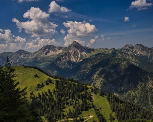A bird's eye view of a mountain range covered with green forests under a cloudy blue sky