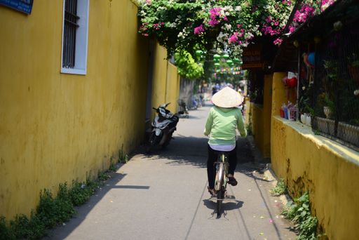 A man riding a bicycle in a narrow street wearing a conical hat
