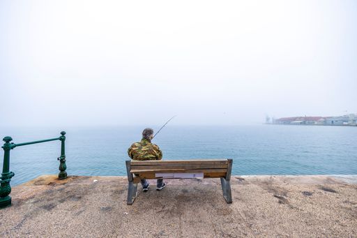A man sitting on a wooden bench, fishing in the sea on a foggy day