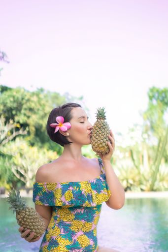 A beautiful caucasian woman with a flower tucked behind her ear holding two fresh pineapples.