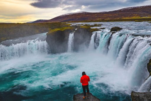 A hiker standing on a cliff near the Godafoos Waterfall under a cloudy sky in Iceland