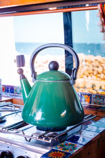 A vertical of a green teapot on the gas in front of the window