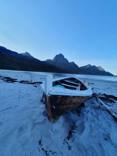 A vertical shot of a small wooden boat covered in snow on the shore with mountains in the background