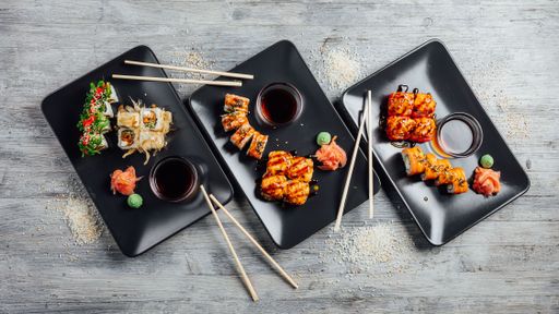 Top view of a set of sushi rolls  on three black plates served with soy sauce, ginger, and wasabi