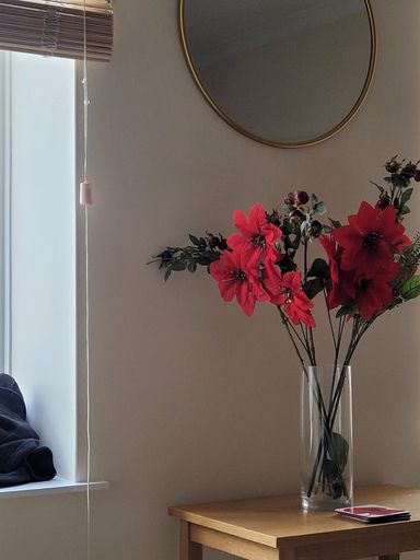 A vertical closeup shot of decorative red flowers in a glass vase on a wooden table beside a window
