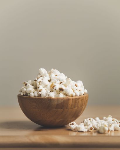 A wooden bowl containing freshly-popped popcorn, with two pieces of the popped kernels on the table