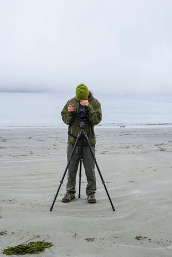 A vertical of a photographer standing in front of the camera on a tripod at the sandy beach on a misty day in Tofino, British Columbia