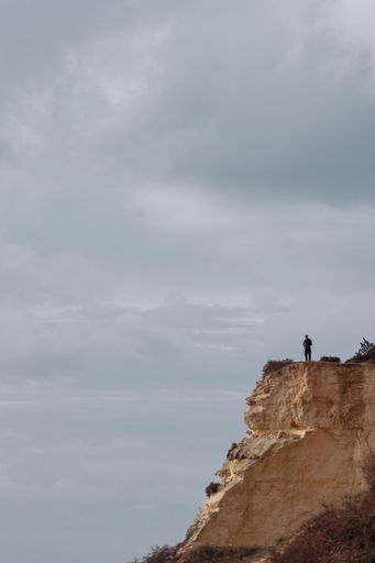 A vertical shot of a man standing on a cliff against the cloudy sky in Algarve, Portugal