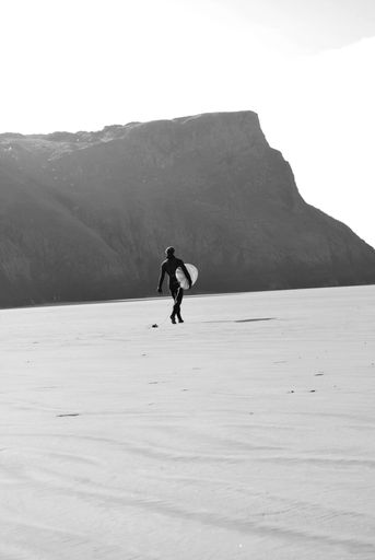 A vertical monochrome shot of a surfer walking along a sandy coast with a board