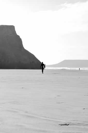 A vertical monochrome shot of a surfer walking along a sandy coast with a board