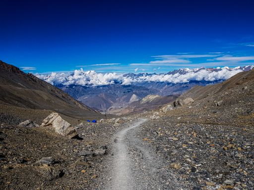 A rocky mountain trail looking at the peaks of a mountain range covered by fluffy clouds