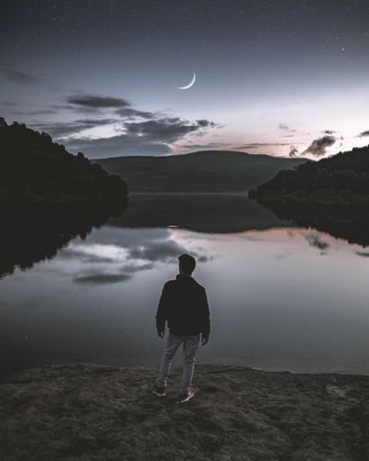 Man in black jacket and red shoes standing in front of a lake and a sunset sky with a moon during du