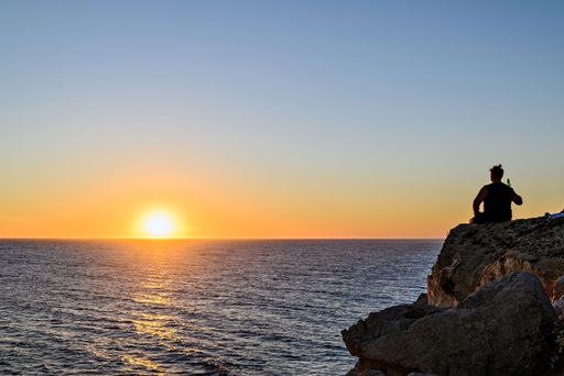 A young man sitting on the rock is drinking a beer and watching the sunset from the sea.