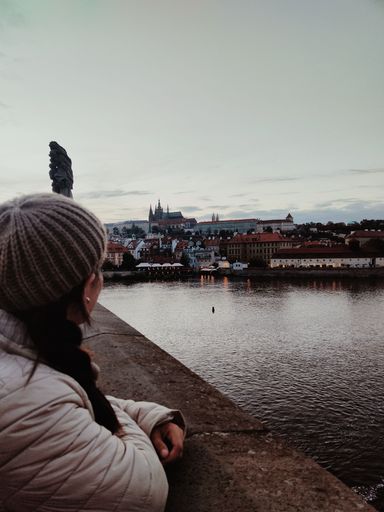 A vertical shot of a woman in a hat looking at Prague city from the Charles Bridge