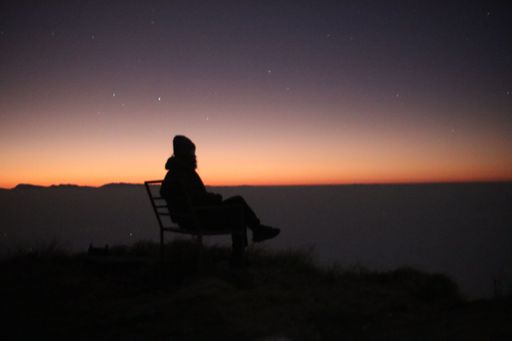 A silhouette of a man sitting on top of a hill, enjoying the view of sunset