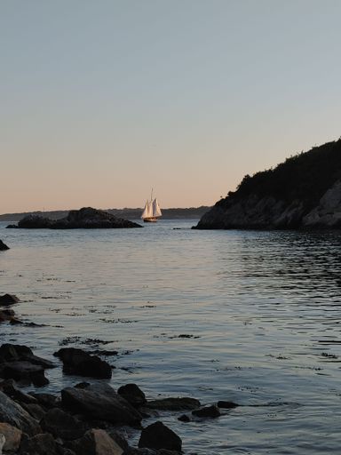 A beautiful seascape of calm waves hitting the rocky shore near the hills and a sailboat floating on background